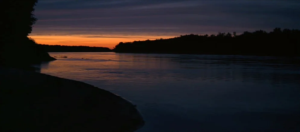 Ride With The Devil movie still: establishing — A wide river winds through a dark landscape under a low-hanging horizon of orange. The water…