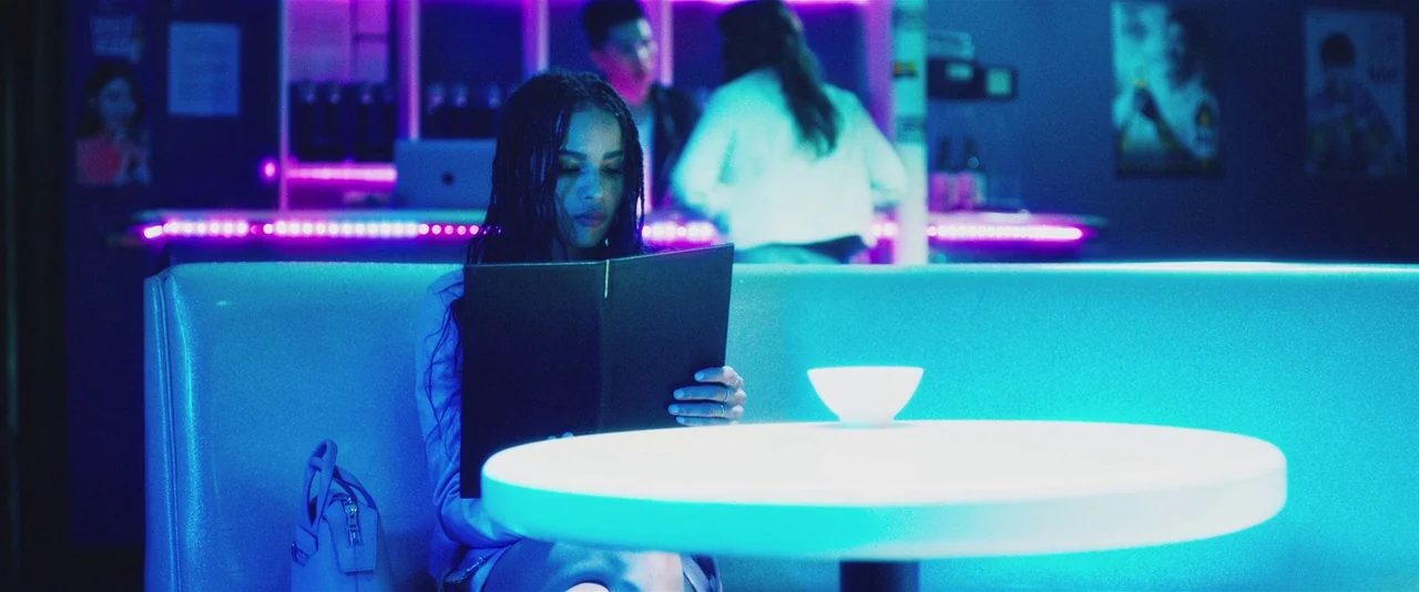 Gemini (2017) movie still: medium — A neon-lit diner interior features a woman sitting alone at a circular table. She is holding a dark menu while…