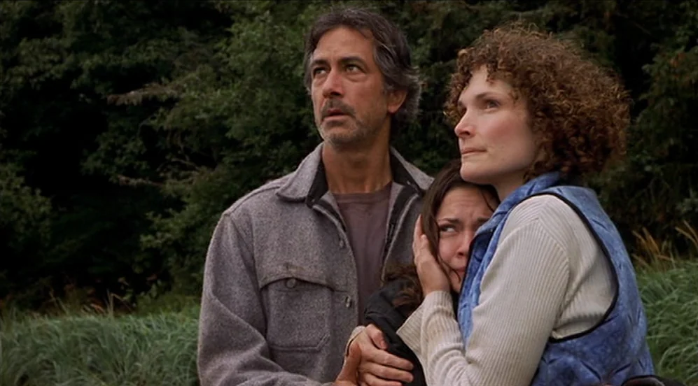 Limbo (1999) movie still: medium — A group of three people stands in a grassy field backed by dark pine trees. They are positioned close together…