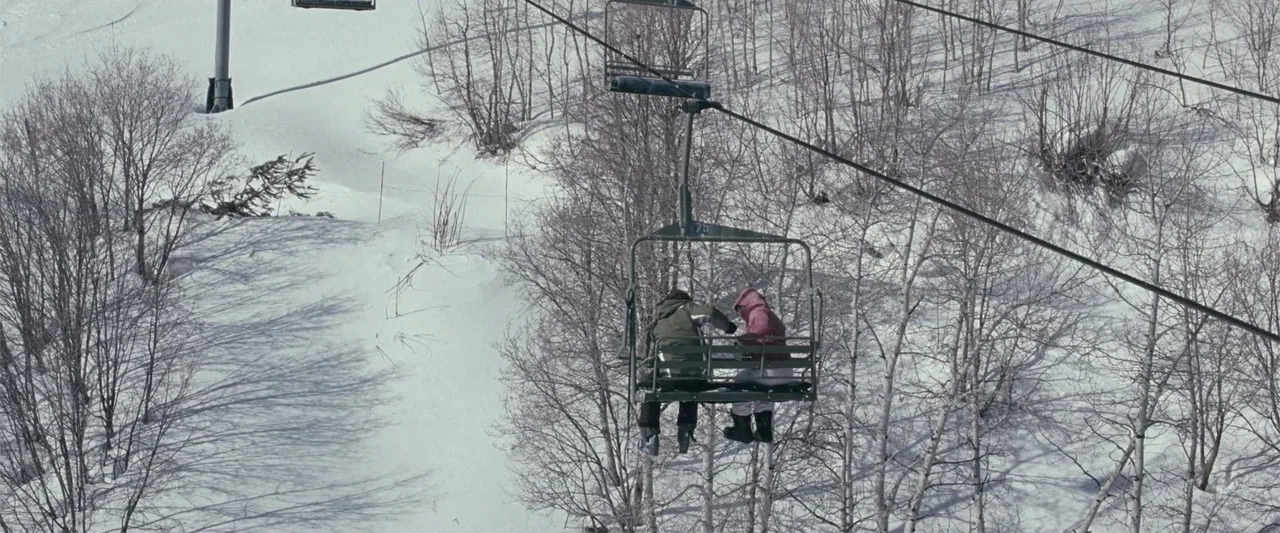 Frozen (2010) movie still: wide — A metal chairlift carries two people over a slope covered in deep white snow. Bare aspen trees grow in dense…