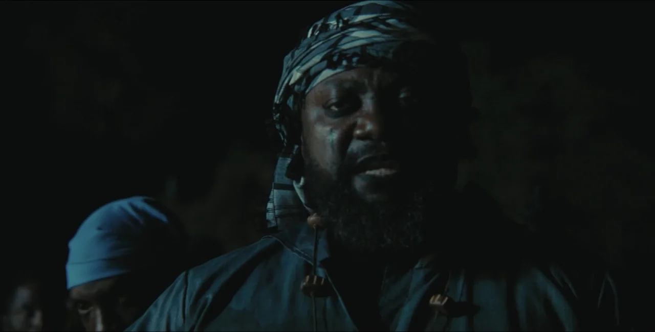 Project Assault (2025) movie still: closeup — A dark outdoor space at night with low-key illumination casting heavy shadows. A man with a patterned…
