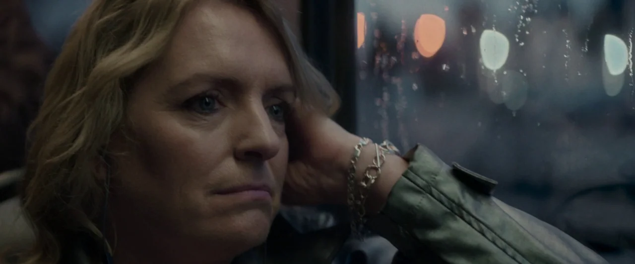 Ali & Ava (2021) movie still: closeup — A woman sits by a wet, raindrop-covered bus window at night in a city. She rests her head on her hand while…