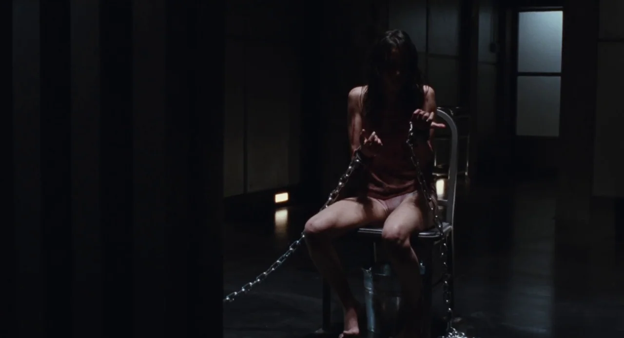 Martyrs (2008) movie still: fullbody — A dark, industrial room with metallic walls and a single metal chair. A woman sits on the chair, her hands…