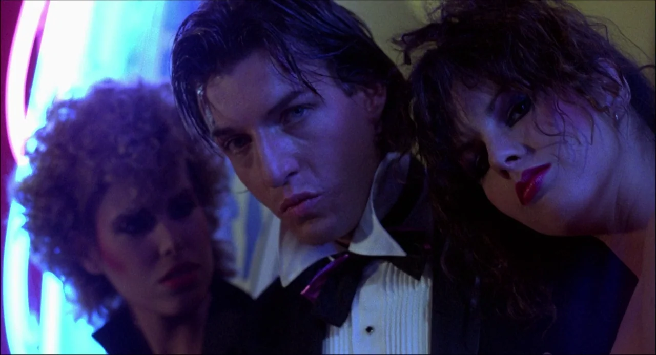 Xanadu (1980) movie still: closeup — A dimly lit space with bright pink and blue neon lights in the background. A man in a tuxedo is flanked by two…