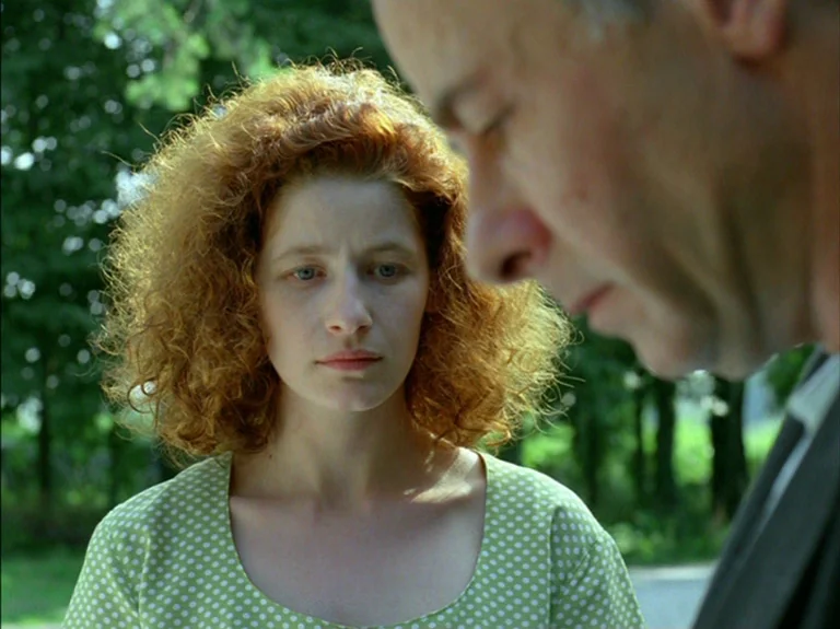Helas Pour Moi (1993) movie still: medium — A woman stands in a tree-lined outdoor space with green foliage in the background. She looks toward a…