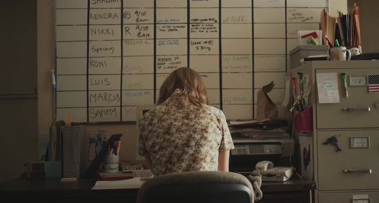 Short Term 12 (2013) movie still: medium — A cluttered nurse's station features a large wall-mounted dry-erase board covered in schedules. A woman…