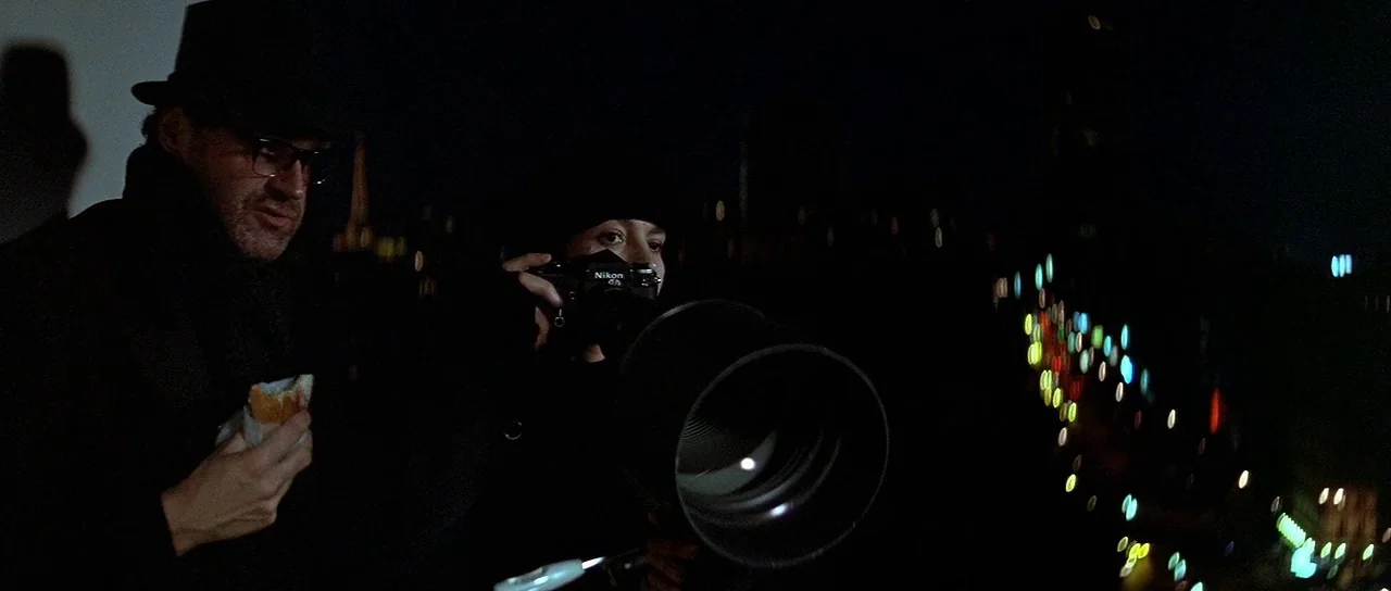 Nikita (1990) movie still: medium — A dark rooftop terrace at night overlooks an out-of-focus city street below. A man eats a sandwich while beside…