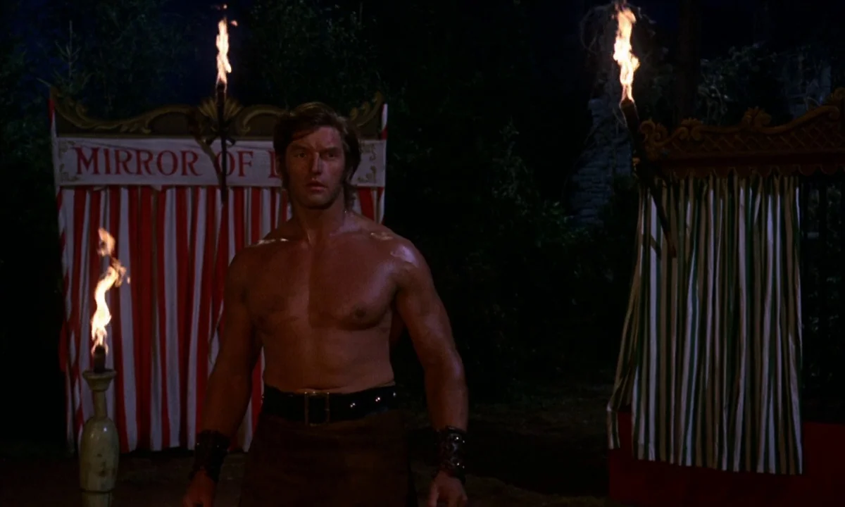 Vampire Circus (1972) movie still: medium — A night scene featuring a carnival tent with striped curtains and a sign. A muscular man stands topless…