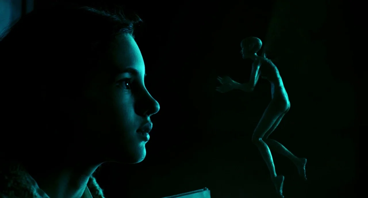 Pan’s Labyrinth (2006) movie still: closeup — A dark, enclosed space is lit by a dim source of cyan illumination. A young girl gazes toward a small,…