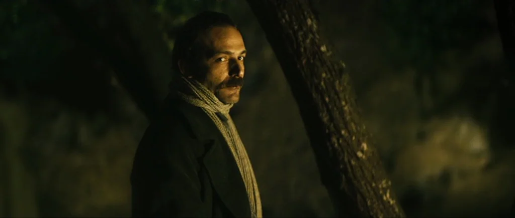 Once Upon a Time in Anatolia (2011) movie still: medium — A dimly lit outdoor space at night with a tree trunk visible in the foreground. A man…