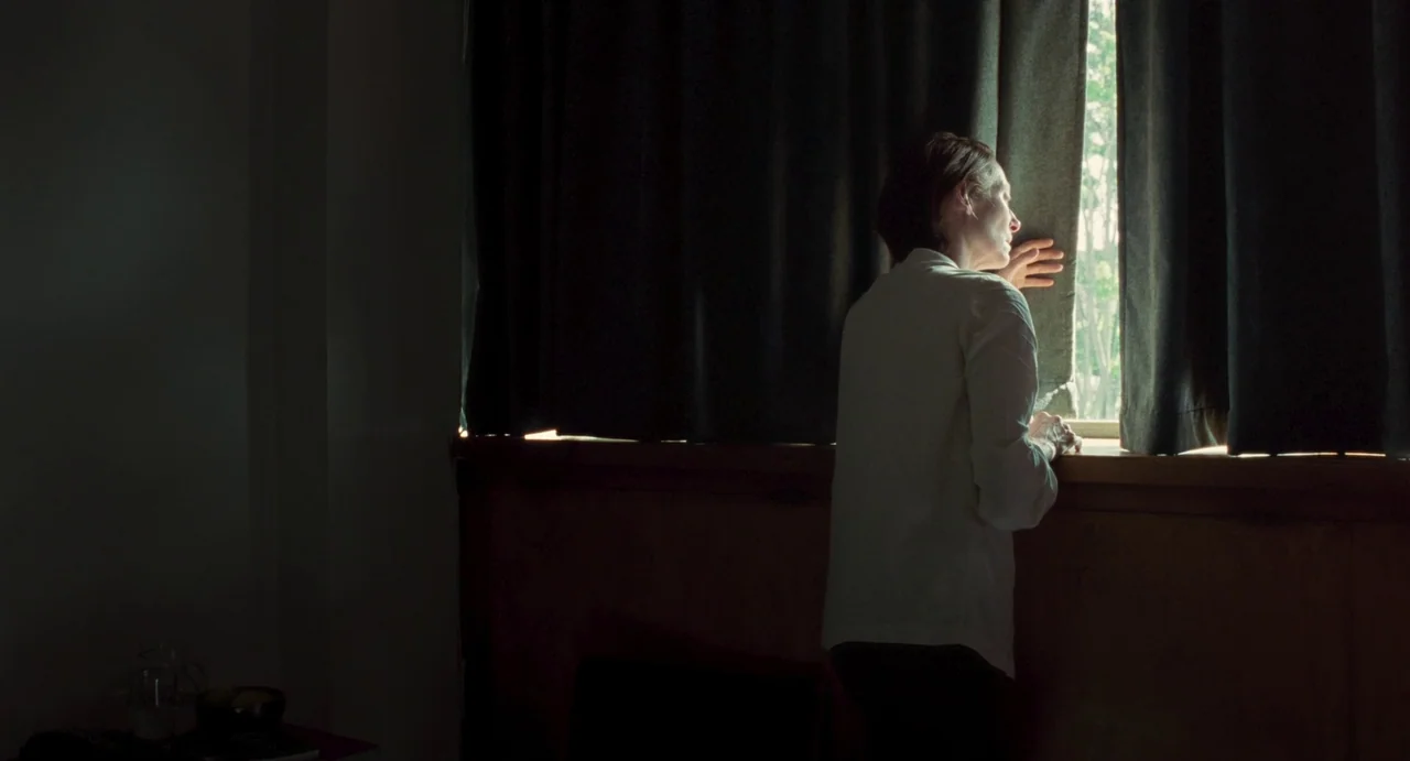 Memoria (2021) movie still: medium — A dark, sparsely furnished room is lit by light coming through a window. A woman in a white shirt stands by the…