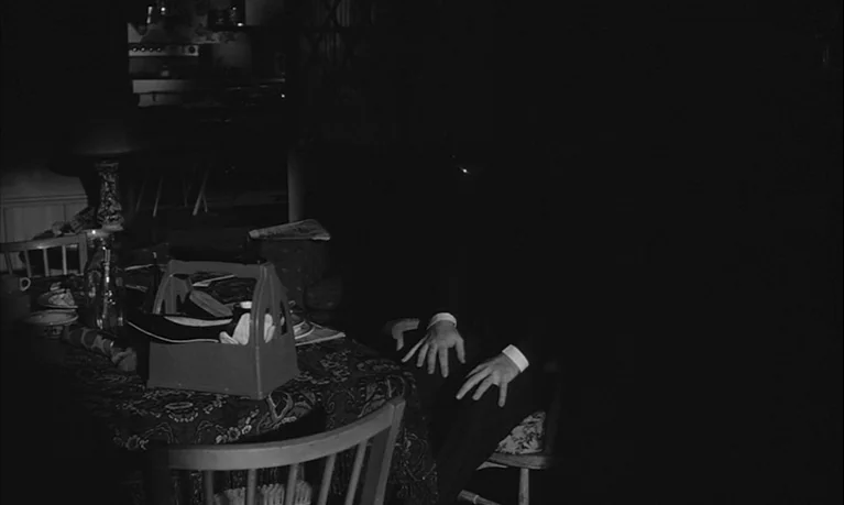 Lolita (1962) movie still: medium — A dimly lit room features a dining table covered by a patterned cloth. A person in a suit sits at the table with…