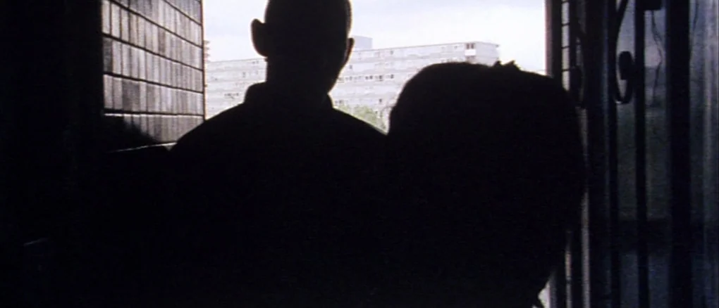 Wonderland (1999) movie still: medium — A dark indoor doorway frames two figures looking out at a residential apartment complex. The bright daylight…
