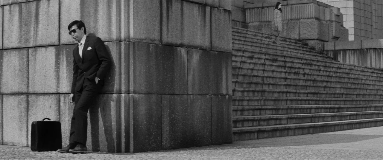 Branded To Kill (1967) movie still: wide — A massive stone monument with wide stairs stands against a clear bright sky. A man waits with a briefcase…