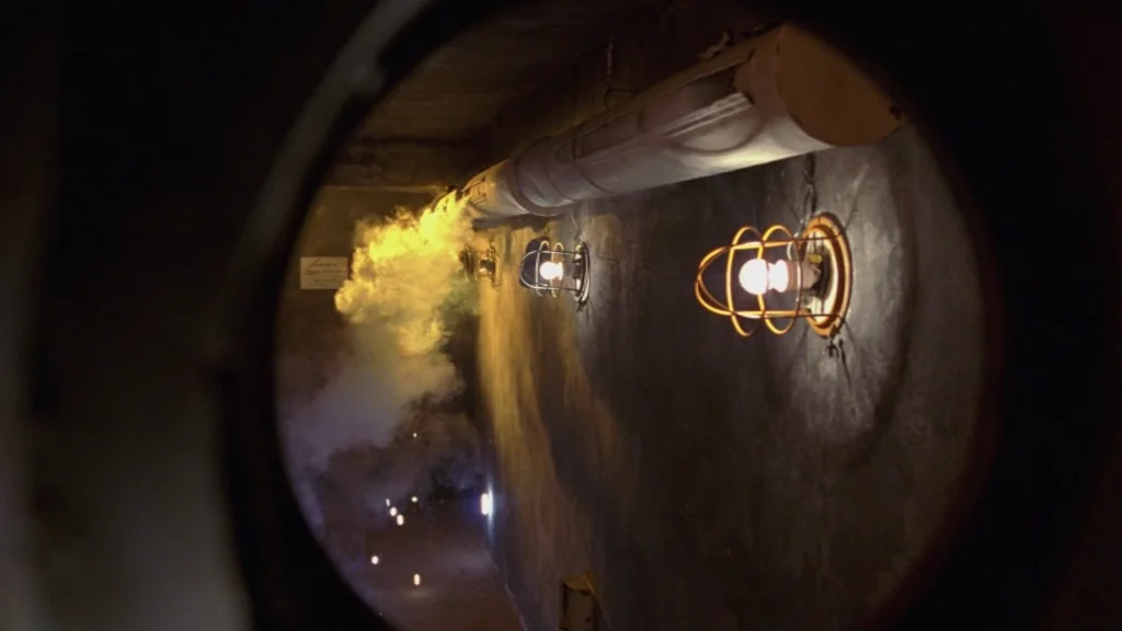 The Ugly Swans (2006) movie still: establishing — A narrow concrete hallway features industrial cage lights mounted along the wall. Smoke pours from…