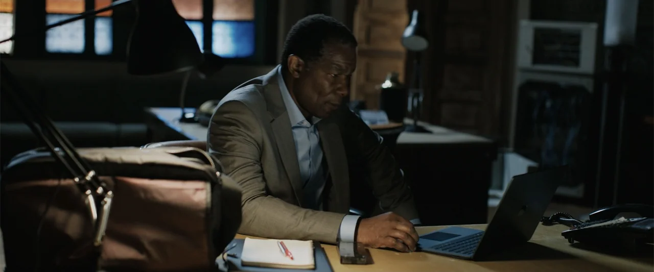 The 355 (2022) movie still: medium — A dimly lit professional office space contains a desk, computer equipment, and paper documents. A mature man in…