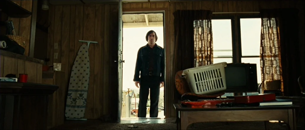 No Country For Old Men movie still: wide — An aged trailer interior features wood-paneled walls and dated patterned curtains. A man stands perfectly…