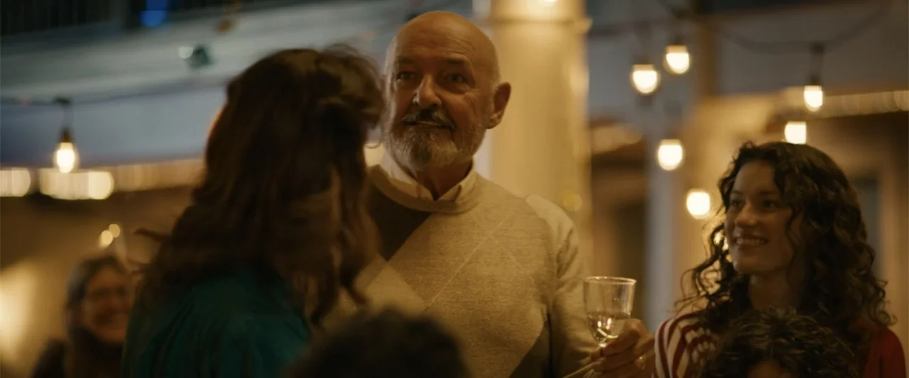 Unsung Hero (2024) movie still: medium — An outdoor social gathering space at night with string lights hung overhead. An older man in an argyle…
