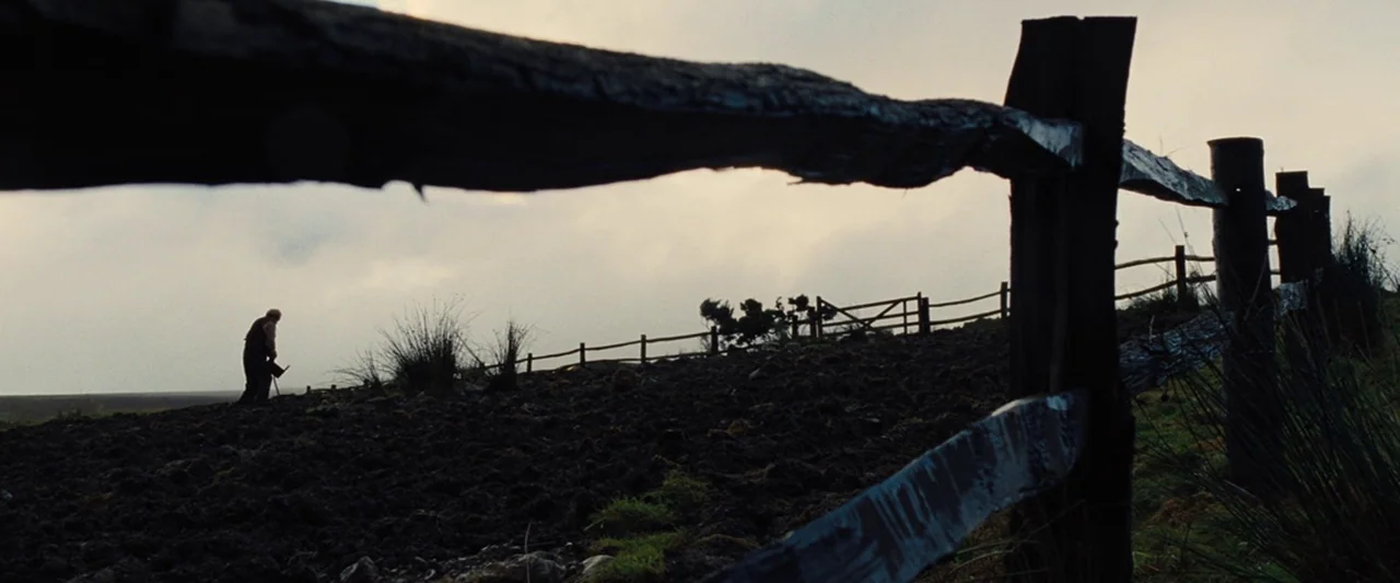 War Horse (2011) movie still: wide — A dark, uneven field is bordered by a rustic wooden fence under a cloudy sky. An elderly man walks alone…