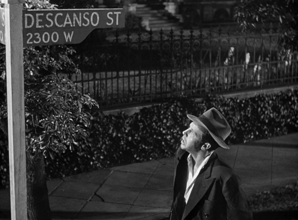 Murder, My Sweet (1944) movie still: medium — A night street corner in a residential area featuring a prominent street sign. A man stands on the…