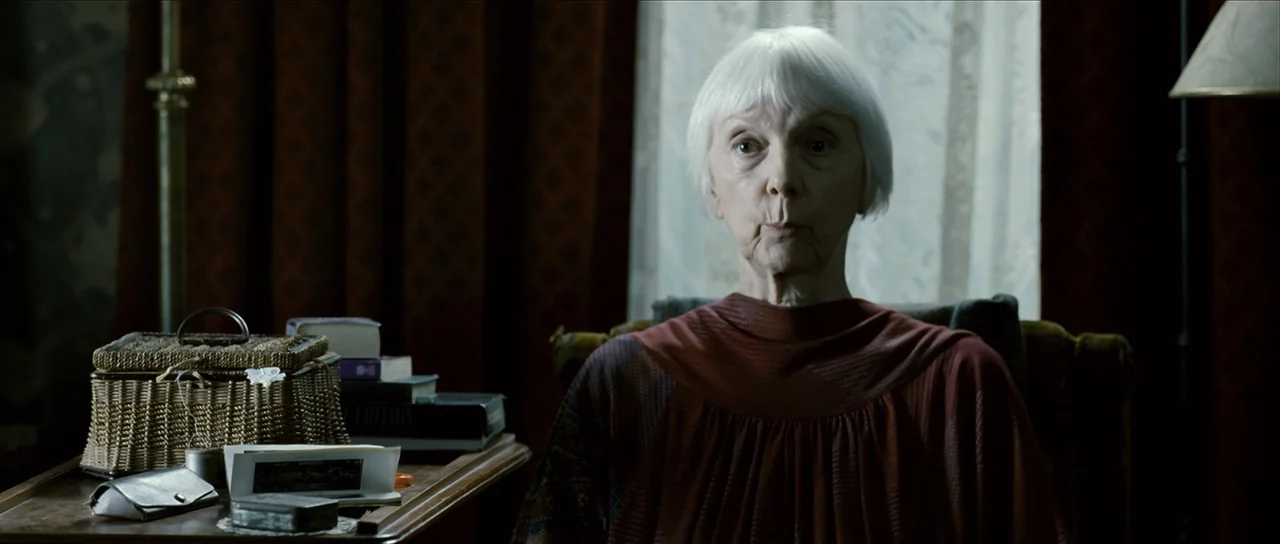 The Oxford Murders (2008) movie still: medium — An interior space features dark red velvet curtains and a wooden table with accessories. An elderly…