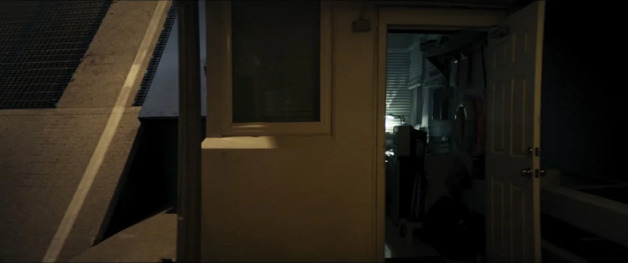 Offseason (2022) movie still: wide — An open doorway reveals the cluttered interior of a small lighted security office at night. The exterior…