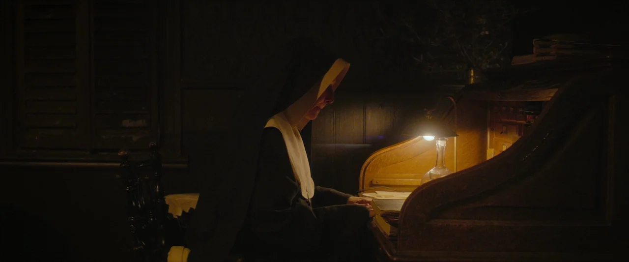 The New Boy (2023) movie still: medium — A dim room contains a wooden roll-top desk illuminated by a small lamp. A nun sits at the desk while…