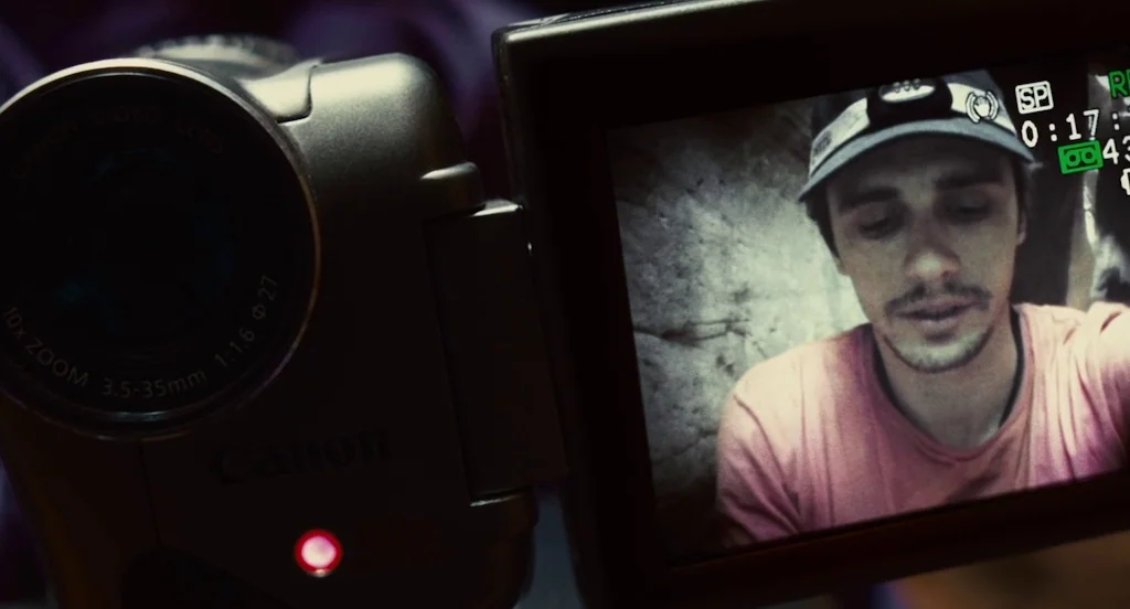 127 Hours (2010) movie still: closeup — A close-up of a consumer-grade Canon camcorder showing a man's face on the screen. The man wears a headlamp…