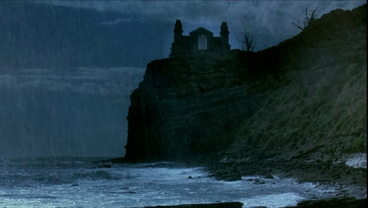 Necronomicon: Book of Dead (1993) movie still: establishing — A dark stone structure stands atop a steep jagged cliff overlooking a churning ocean.…