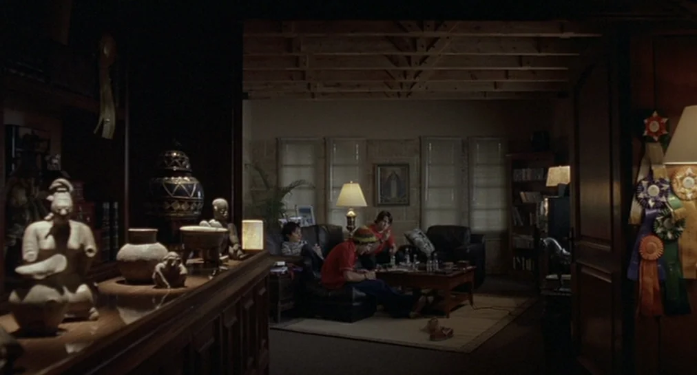 Y Tu Mamá También (2001) movie still: wide — A dimly lit room viewed from an adjacent space across a wooden console table. Three young men sit on a…