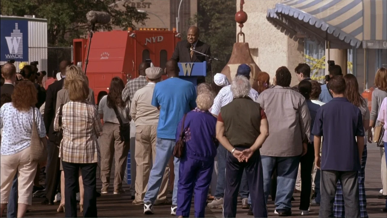 Two Weeks Notice (2002) movie still: medium — A diverse crowd of people stands on a wooden boardwalk at a public event. An elderly man in a suit…