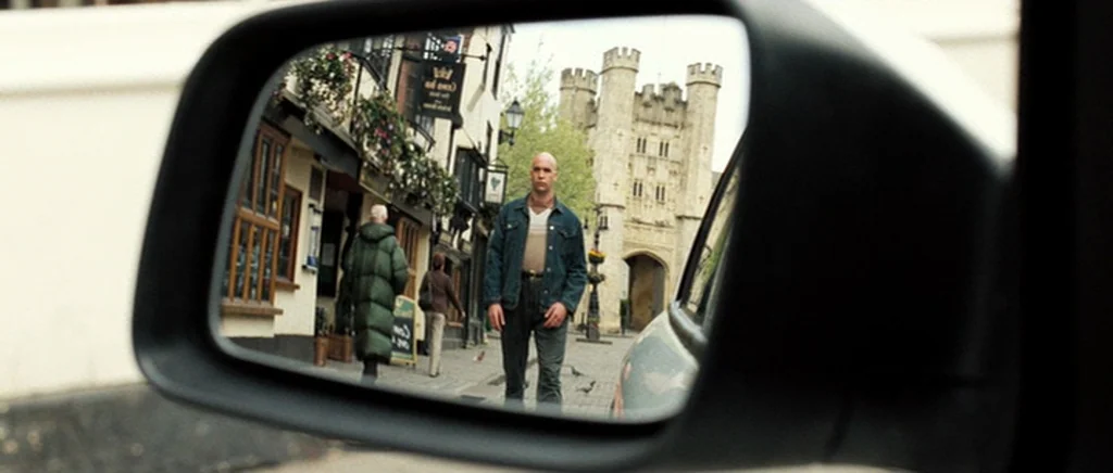 Hot Fuzz (2007) movie still: closeup — An English historic street with a stone gateway is viewed through a car mirror. A bald man wearing a denim…