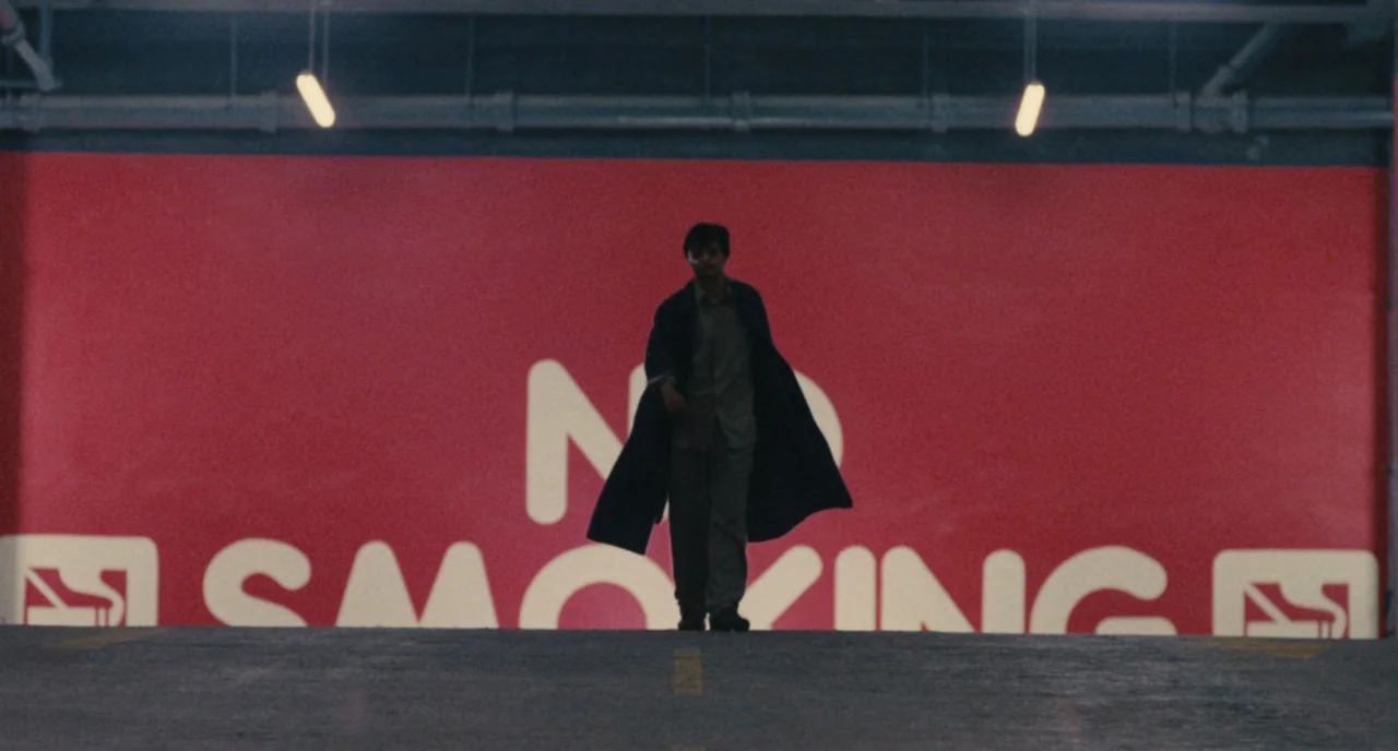 A Better Tomorrow (1986) movie still: wide — A man walks forward in a parking structure with a large red wall behind him. The wall features massive…