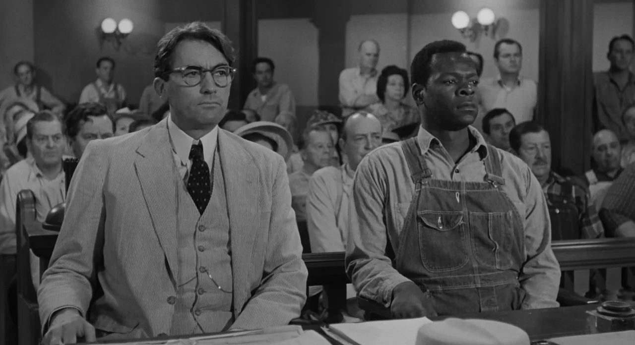 To Kill A Mockingbird (1962) movie still: medium — A crowded courtroom features wood paneling and multiple rows of observers sitting in benches. Two…