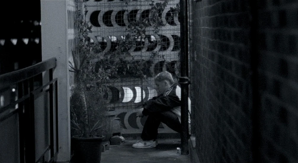 Somers Town (2008) movie still: fullbody — A dark balcony corner features a patterned brick screen wall covered in climbing vines. A teenage boy…