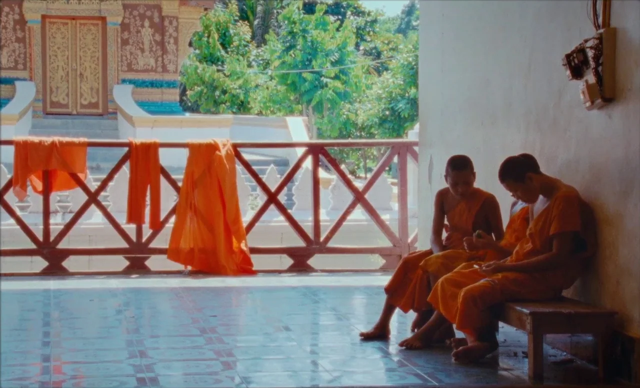 Samsara (2023) movie still: fullbody — A sunny monastery balcony features bright orange robes draped over a simple wooden railing. Two young monks…