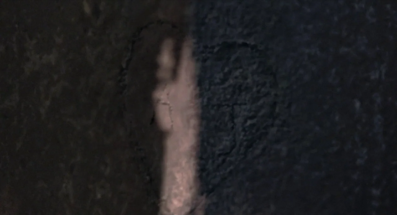 Suspiria (2018) movie still: closeup — A dark, textured wall surface features a subtle, shallow-etched heart shape in the center. A vertical beam of…