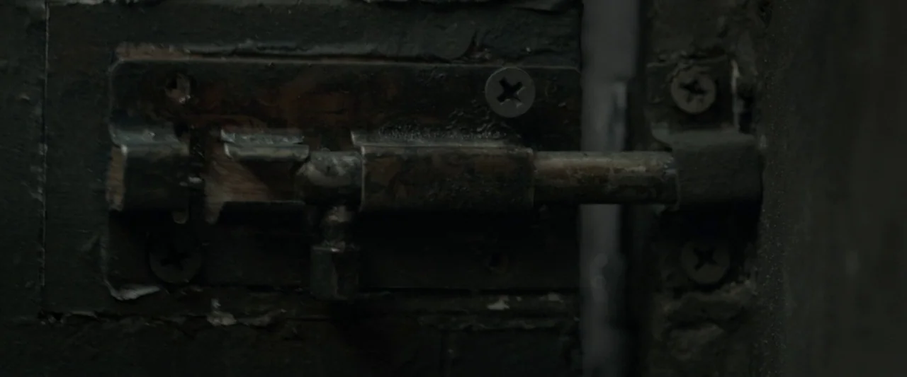 The Raid 2 (2014) movie still: closeup — A close-up of a weathered, dark metal sliding bolt lock mounted on a door. The metal shows signs of rust…