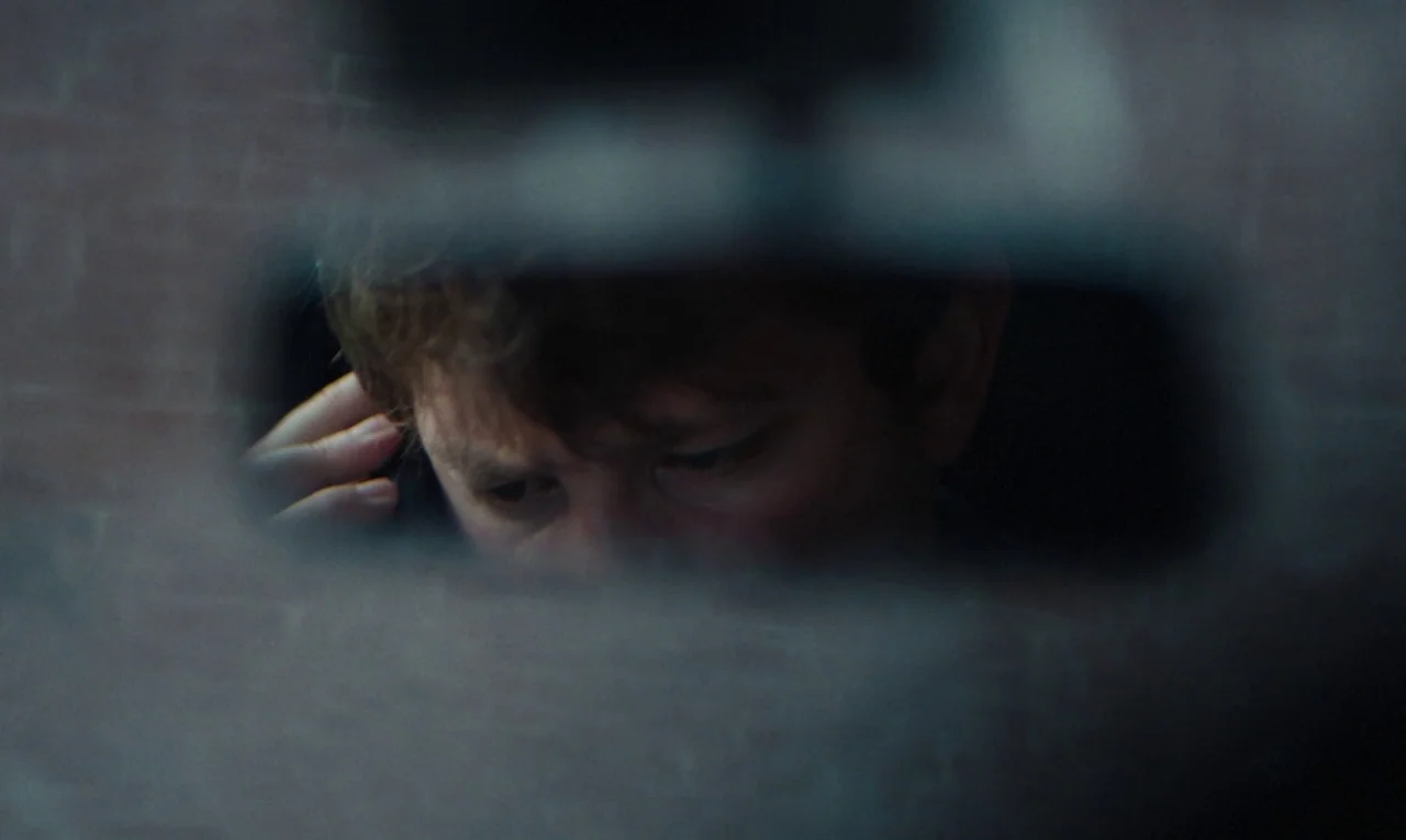 Maldoror (2024) movie still: closeup — A man's face is reflected in a dark rearview mirror inside a vehicle. The subject looks downward toward an…