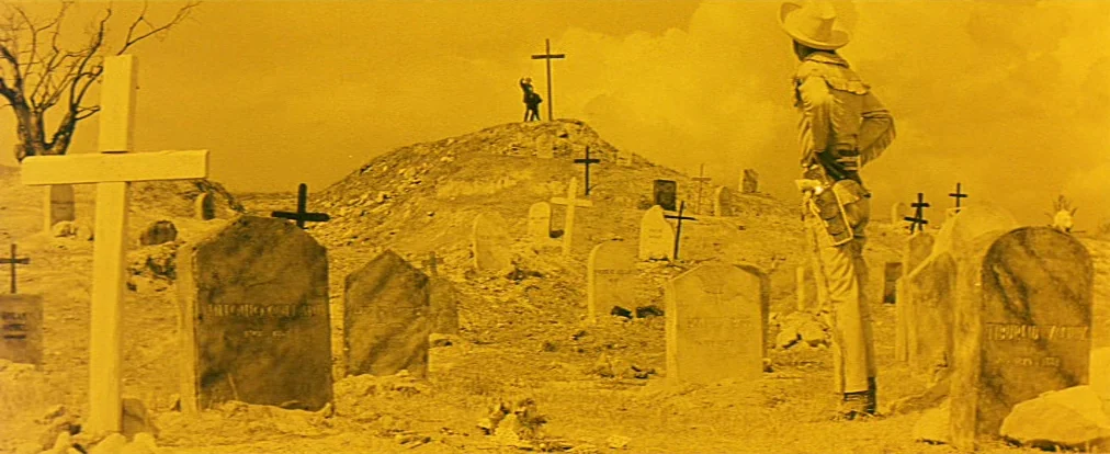 Lemonade Joe (1964) movie still: wide — A dusty, arid cemetery filled with worn headstones and scattered wooden crosses. A man in a cowboy hat…