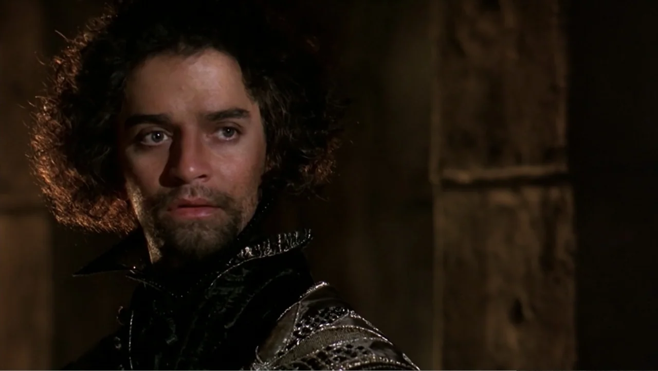 Elizabeth (1998) movie still: closeup — A dimly lit stone interior features a man in period clothing against a shadow. The man with dark curly hair…