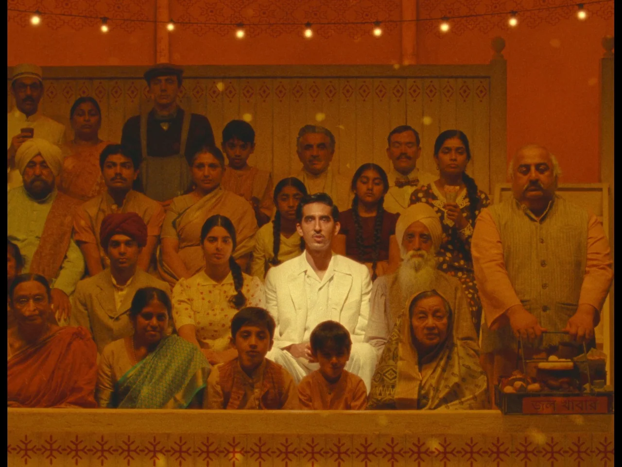 The Wonderful Story of Henry Sugar (2023) movie still: wide — A large group of people poses in an interior space with patterned wooden walls. The…