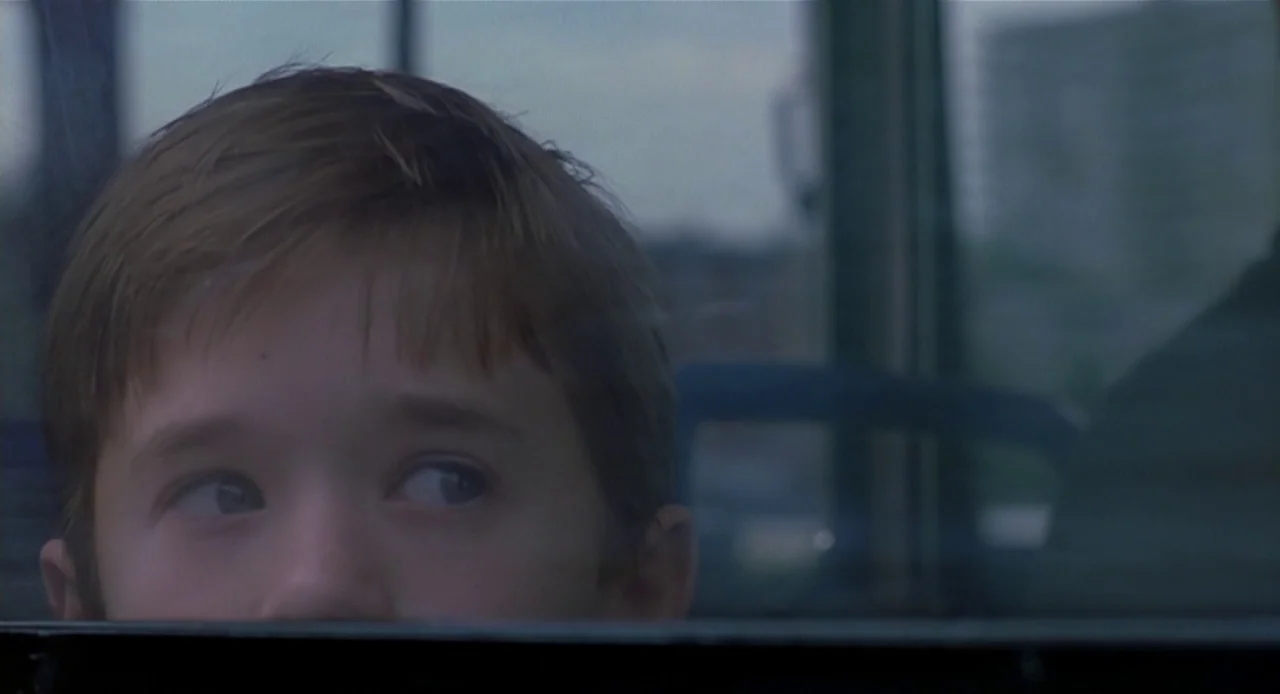 The Sixth Sense (1999) movie still: closeup — A young boy's face is partially visible behind a window frame in a vehicle. The exterior view is…