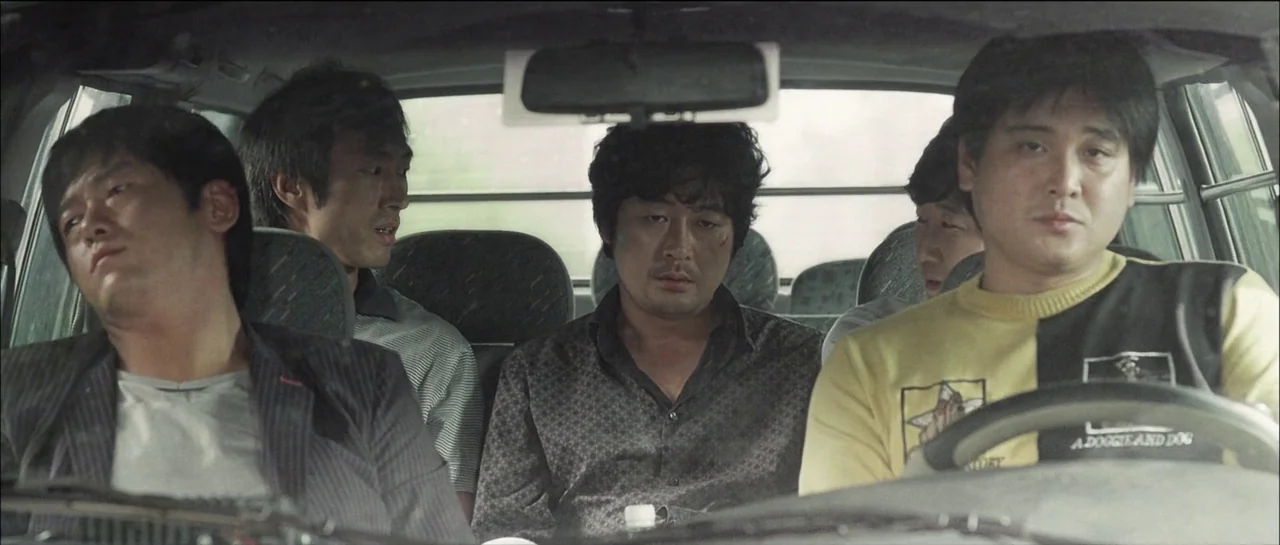 The Chaser (2008) movie still: medium — Four men sit closely together in the front and back seats of a vehicle. They face forward in silence while…