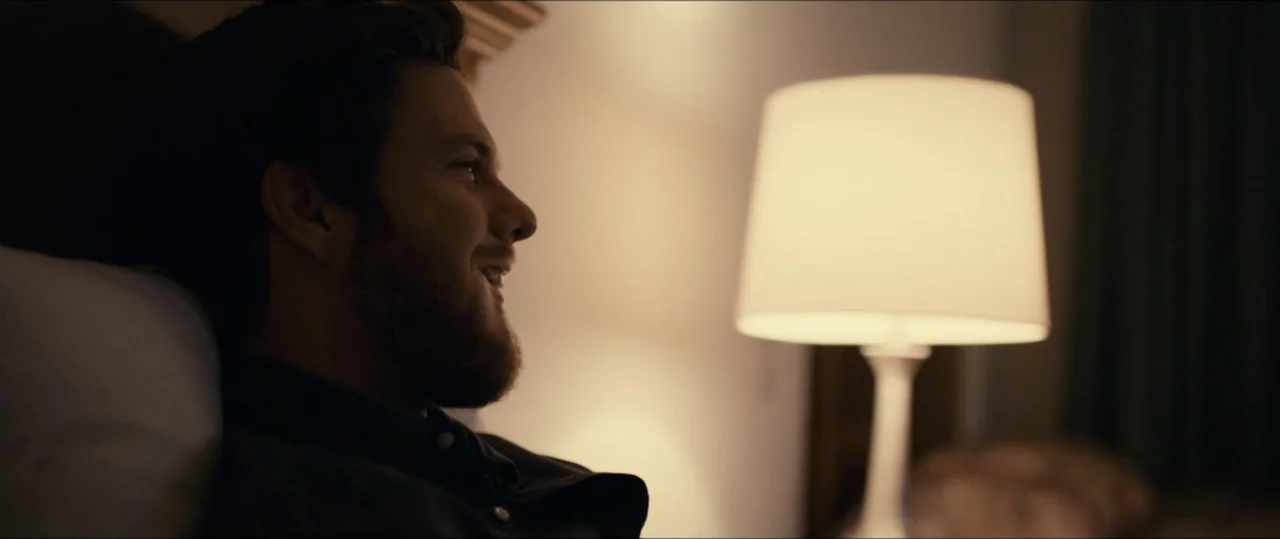 Plus One (2019) movie still: closeup — A dimly lit room contains a person resting in a bed next to a lamp. A bearded man in a dark shirt lies…