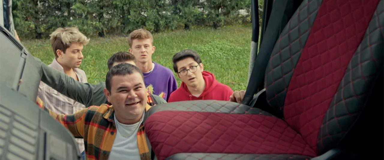 Wrong Package (2025) movie still: medium — A group of four young men look through a car door window from outside. The car interior features quilted…