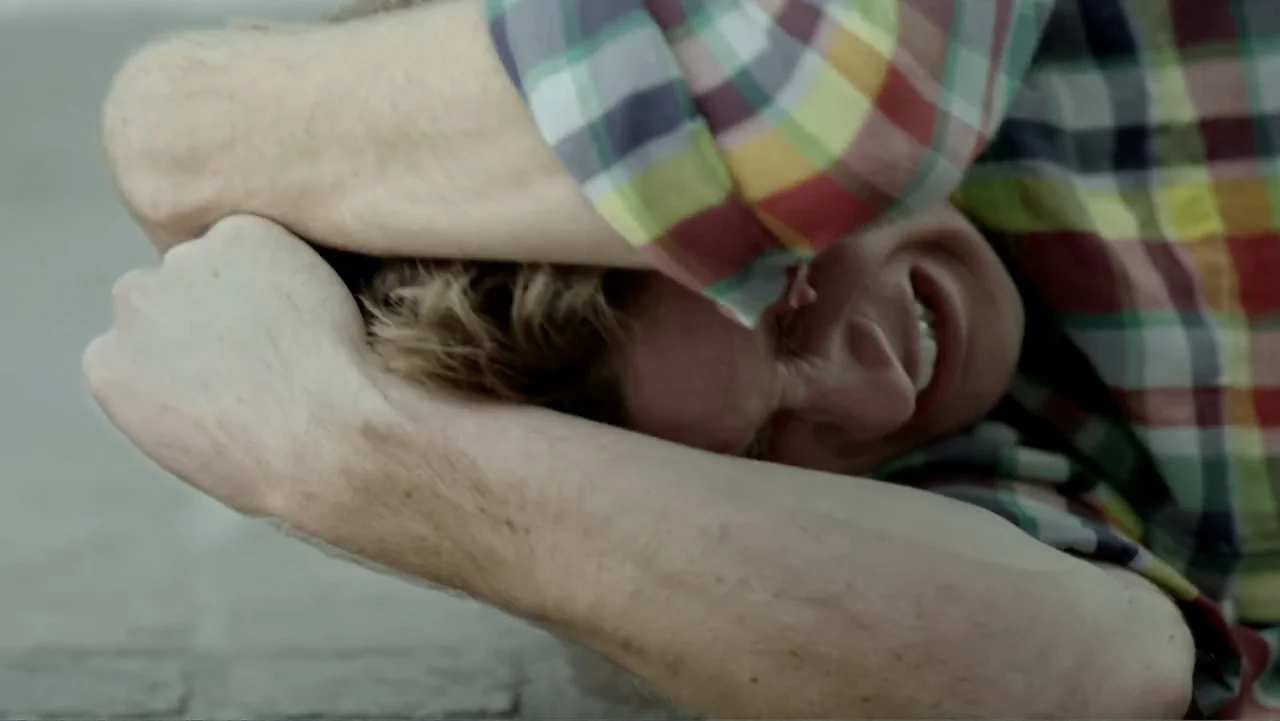 Loco Love (2003) movie still: closeup — A man lies prone on grey outdoor concrete ground in a public space. He curls his body tightly with both arms…