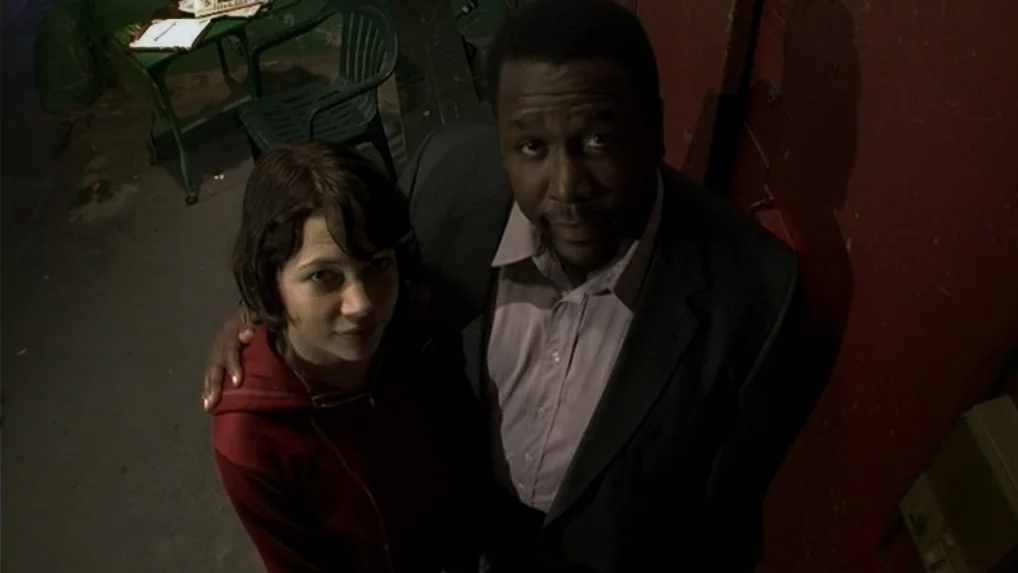 Land of Plenty (2004) movie still: medium — An outdoor paved area with a red painted wall and a green plastic chair. Two people look up at the…