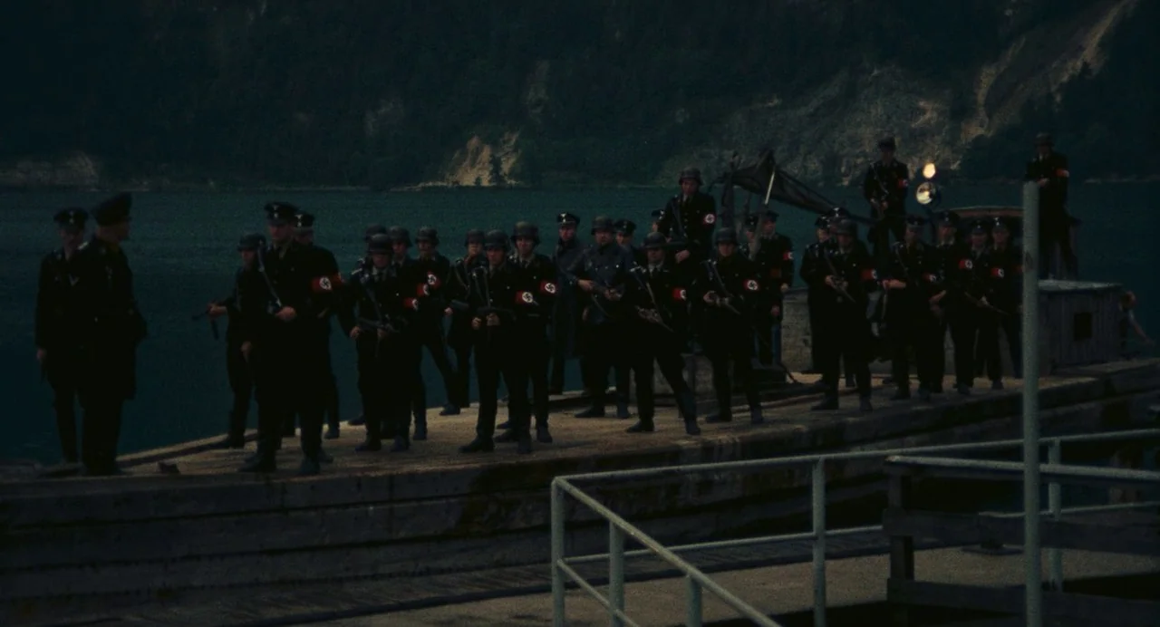 The Damned (1969) movie still: wide — A group of armed soldiers in dark uniforms stands on a concrete pier at dusk. Behind the pier is a dark lake…