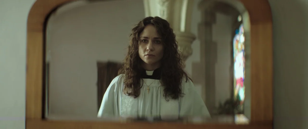 Lord of Misrule (2023) movie still: medium — A woman stands before a mirror in a room with a stained glass window. She stares directly into her own…